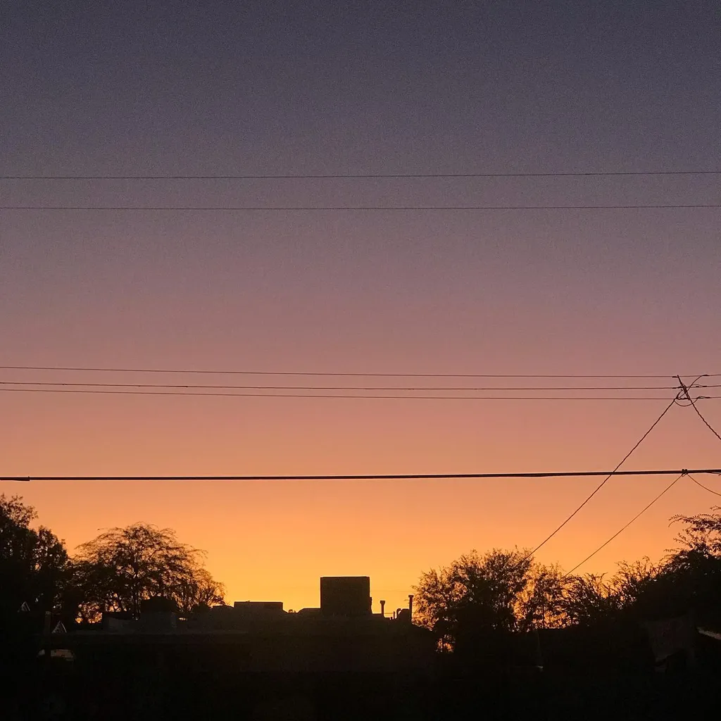 A layered twilight composition featuring a gradient sky from deep indigo to bright orange, above a black silhouette of trees and buildings, intersected by thin horizontal power lines.