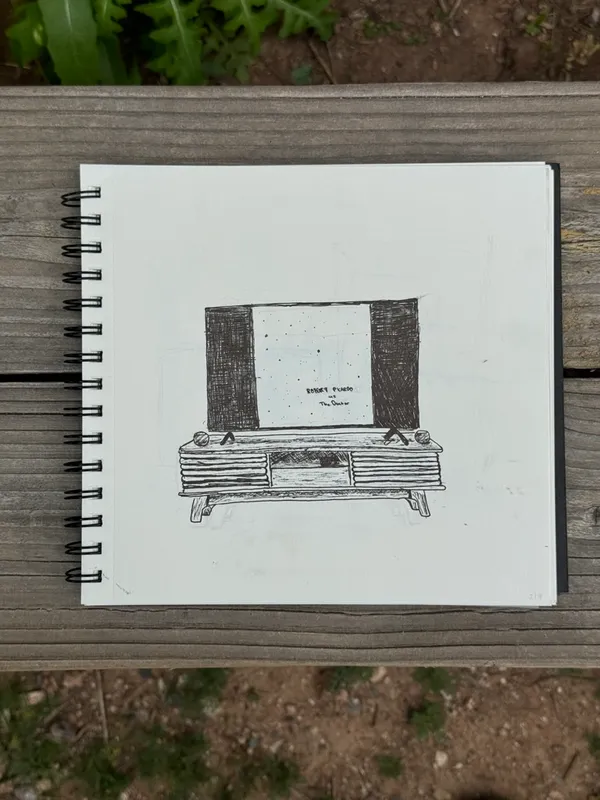 A black and white sketch of a rectangular cabinet with solid black sides and three drawers, drawn on white paper and placed on an open spiral sketchbook on a wooden surface.