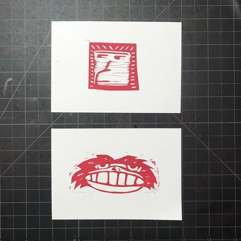 Two red, stylized graphic prints are centered on white paper, featuring a rectangular face design above a wide, aggressive mouth design.
