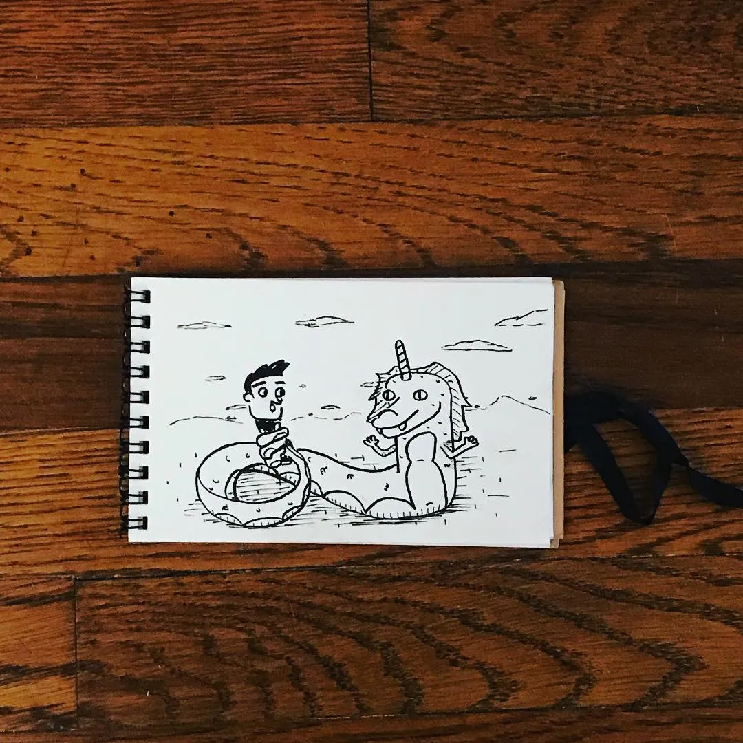 Black ink drawing on white paper depicts a man and a unicorn-like creature sitting together on a sketchbook placed on a brown wooden surface.