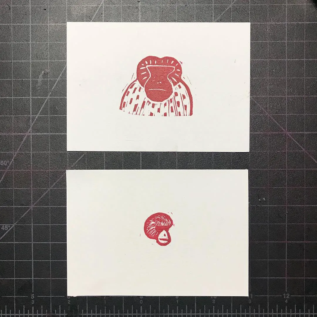 Two red ink drawings of stylized primate faces are presented on white paper, stacked vertically on a grid background. The top drawing is larger and patterned, and the bottom drawing is smaller and solid.