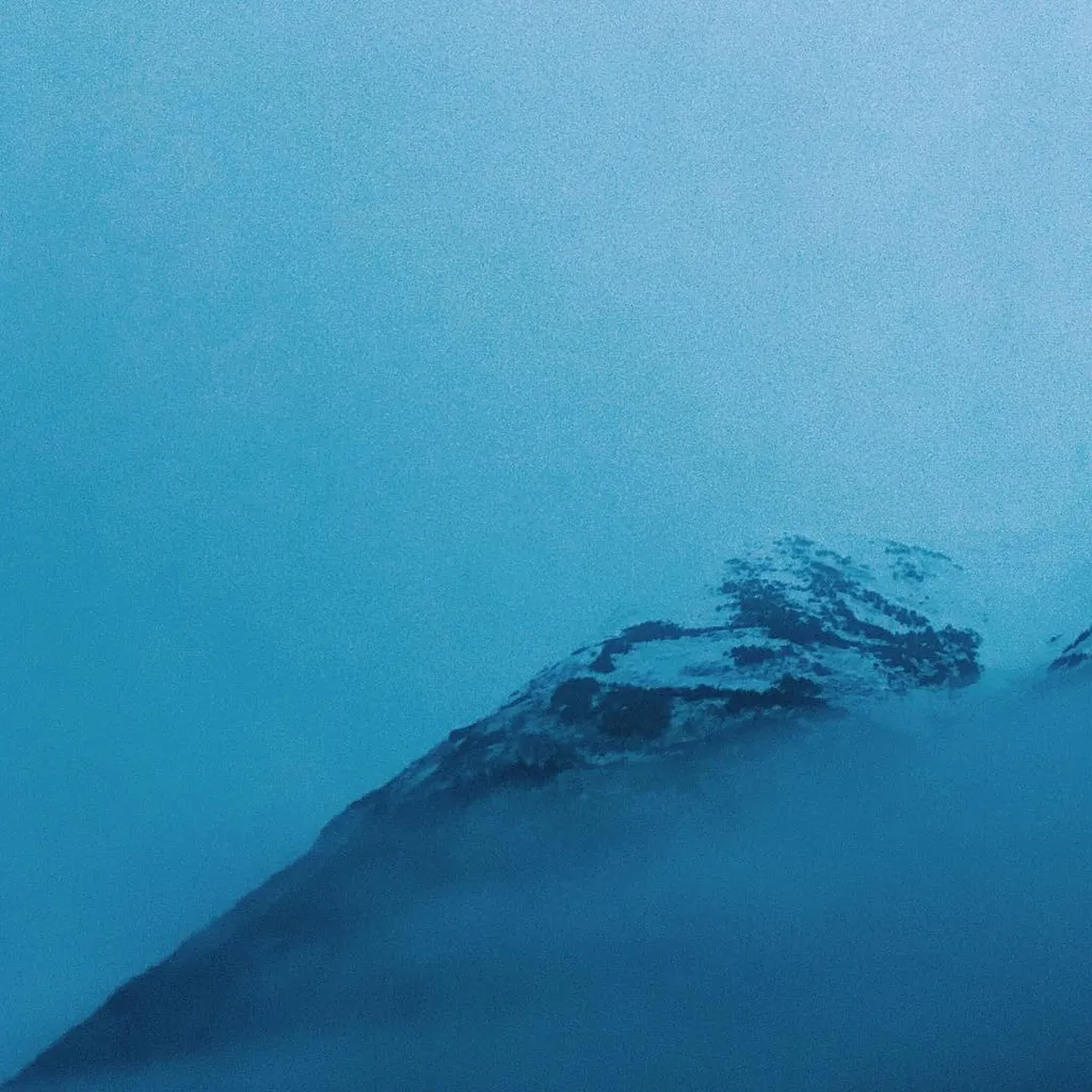 A diagonal mountain ridge emerges from dense blue fog into a lighter, hazy blue sky, dominated by cool, diffused tones.